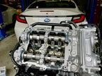 Close-up of a Subaru FA24 engine with Denstoj FA/FB rocker retainers installed beneath the camshafts, showing the assembled valvetrain and cylinder head in a workshop with a Subaru BRZ in the background.