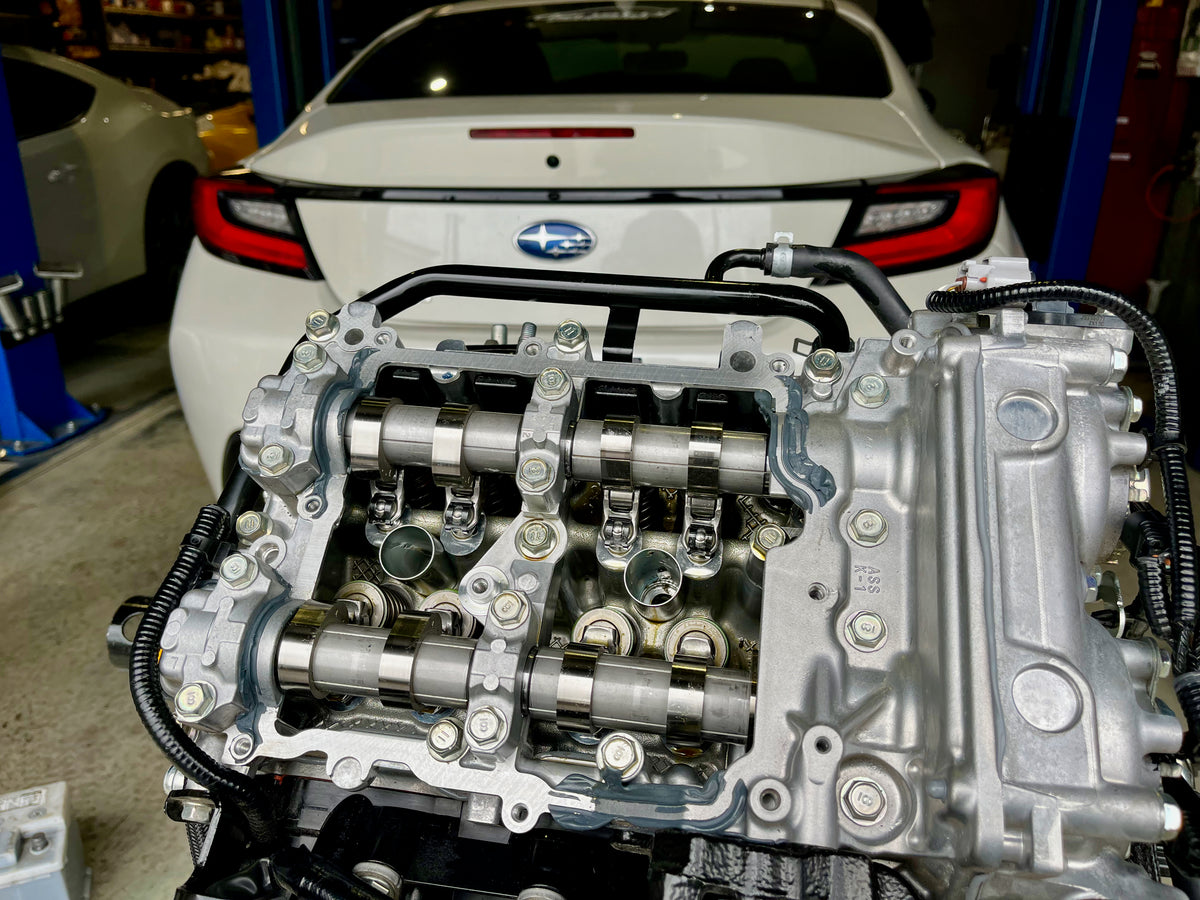 Close-up of a Subaru FA24 engine with Denstoj FA/FB rocker retainers installed beneath the camshafts, showing the assembled valvetrain and cylinder head in a workshop with a Subaru BRZ in the background.